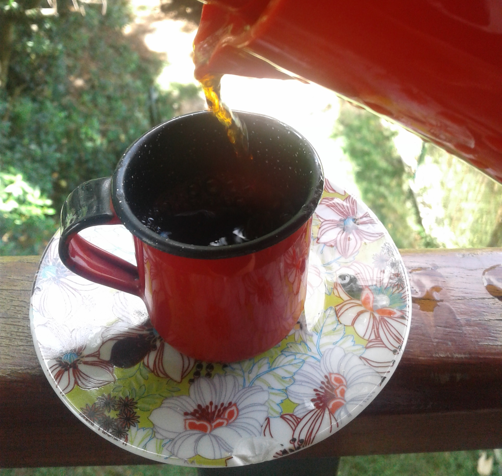 #paracegover : café sendo servido de uma cafeteira vermelha, em uma xícara vermelha de ágata, com pratinho colorido; ao fundo, desfocado, verde da natureza.