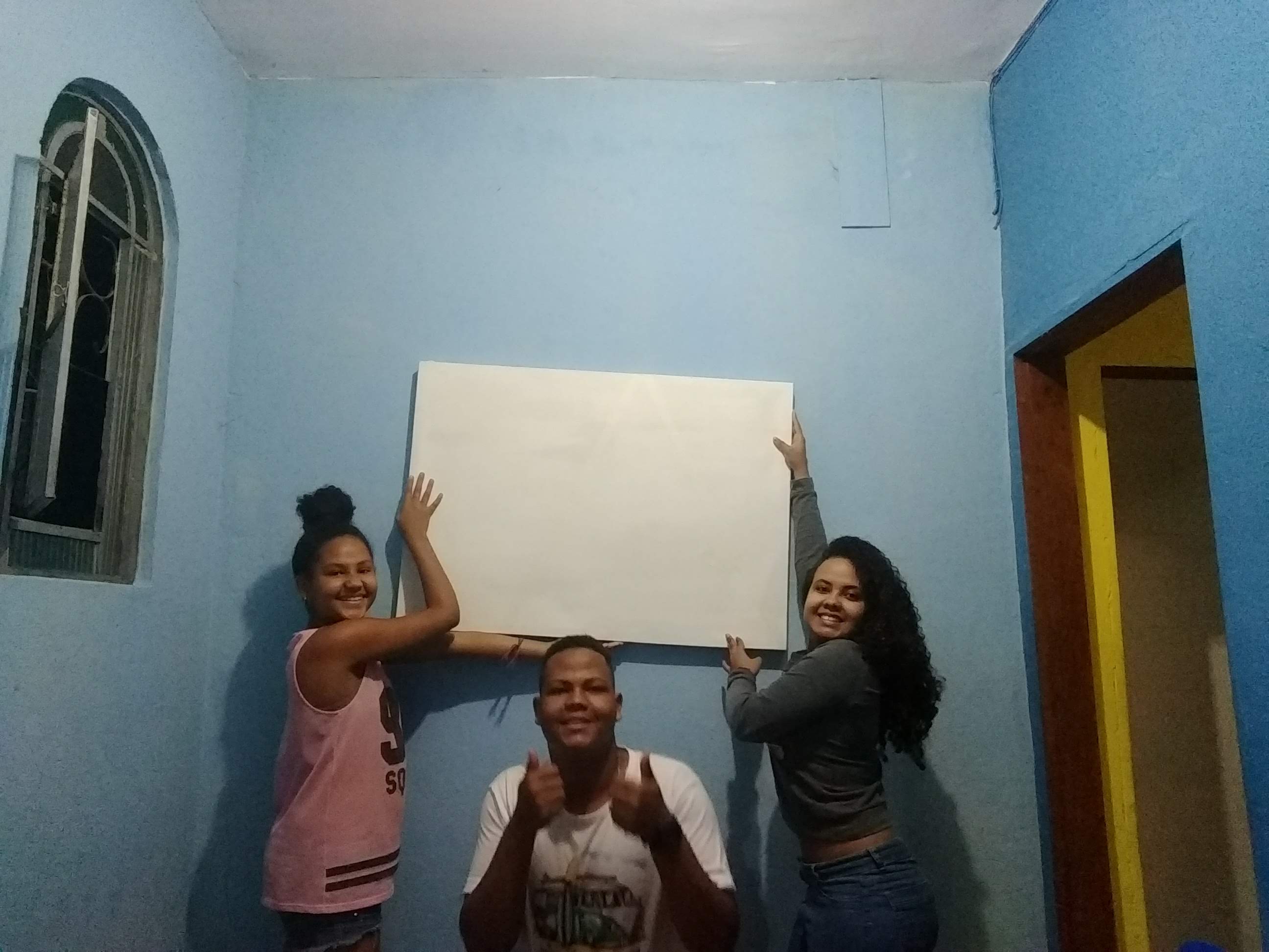 A foto com fundo azul tem três pessoas (duas meninas, uma de cada lado e um menino no centro) segurando uma tela de pintura que está centralizada. A tela está em branco. Cada um faz uma pose e tem no rosto uma expressão de alegria.