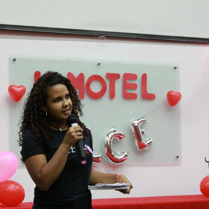 Jady, cabelos cacheados com um microfone. Falando em um palco com decoração vermelha no fundo. Ela usa uma blusa da faculdade de letras